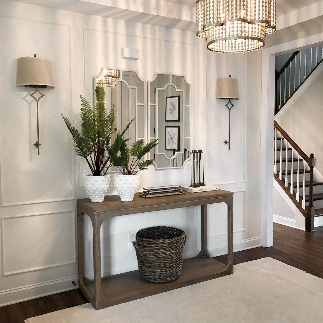 A console table holds potted ferns and decorative items, reflecting light from a chandelier while a staircase ascends in the background of a stylish entryway.