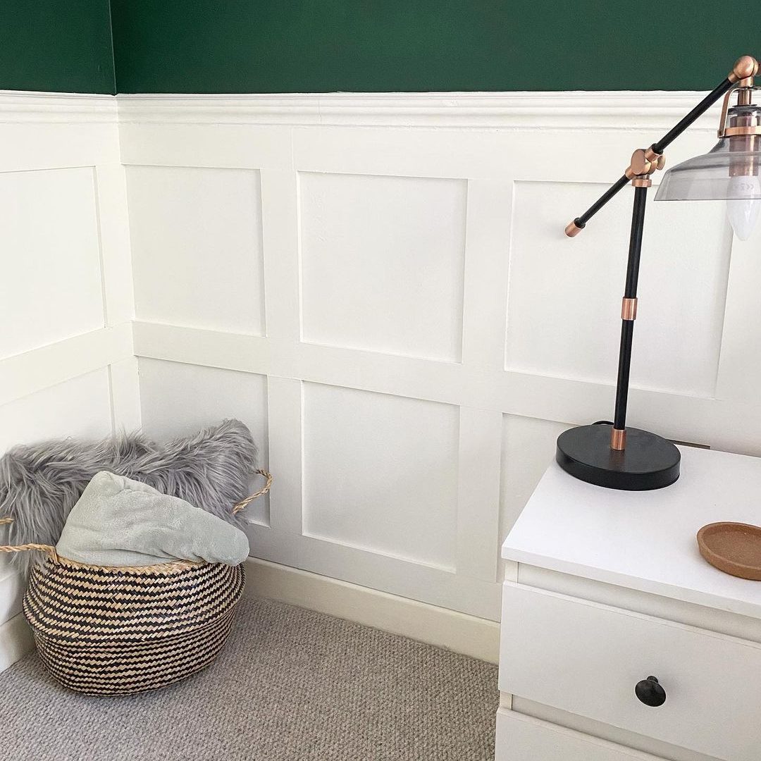 A black and copper desk lamp illuminates a cozy corner with a woven basket holding pillows, near a white nightstand against green walls.