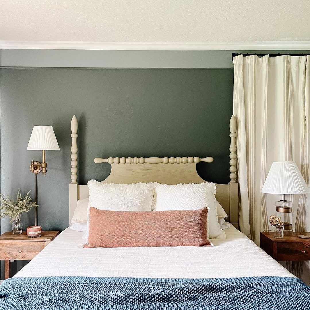A light-colored bed with decorative pillows sits against a dark wall, flanked by lamps and a small nightstand, creating a cozy bedroom atmosphere.