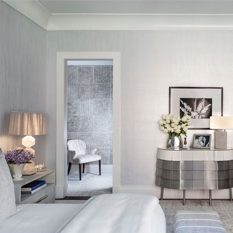 A bedside lamp illuminates a white bed; a modern table displays flowers and photos, leading to a doorway revealing a simple chair in a textured room.