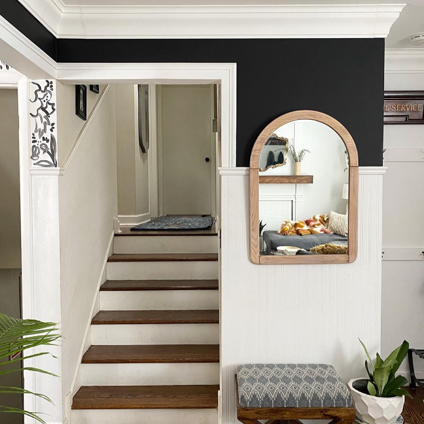 A wooden-framed mirror reflects a cozy living space, while stairs ascend to a door under a neatly painted wall with decorative accents.