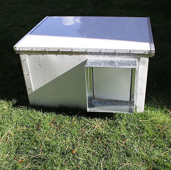 A silver metal box with a white top sits on green grass, featuring a small access opening on one side, likely for an animal's entry.