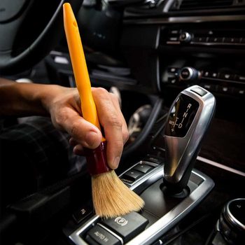 A hand holds a small brush, gently cleaning the gear shift area inside a car, surrounded by black interior and various controls.