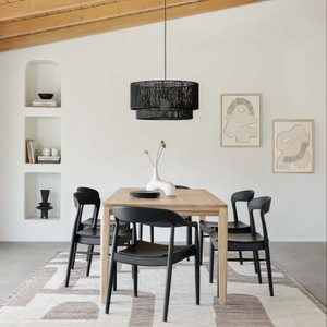 A wooden dining table with six black chairs sits on a patterned rug. Above hangs a pendant light, complementing minimalist artwork on a white wall.