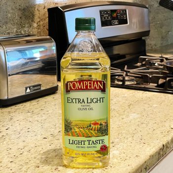 A bottle of Pompeiian extra light olive oil stands on a kitchen counter next to a toaster and a stove with a frying pan.