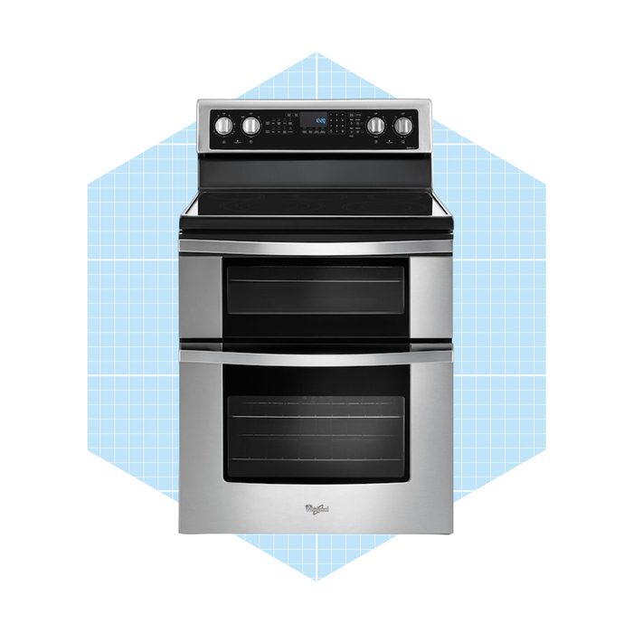 A stainless steel oven with two black glass doors stands against a light blue geometric background, displaying control knobs and a digital display at the top.