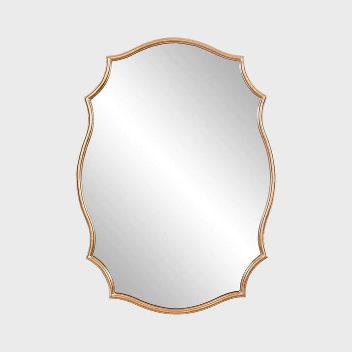 An ornate, gold-framed mirror reflects light, hanging alone against a neutral background, adding elegance and depth to its surroundings.