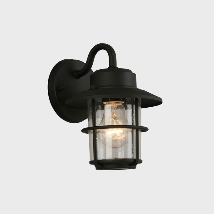 A black outdoor wall lantern illuminates a clear glass enclosure, featuring a visible light bulb inside, mounted against a neutral background.