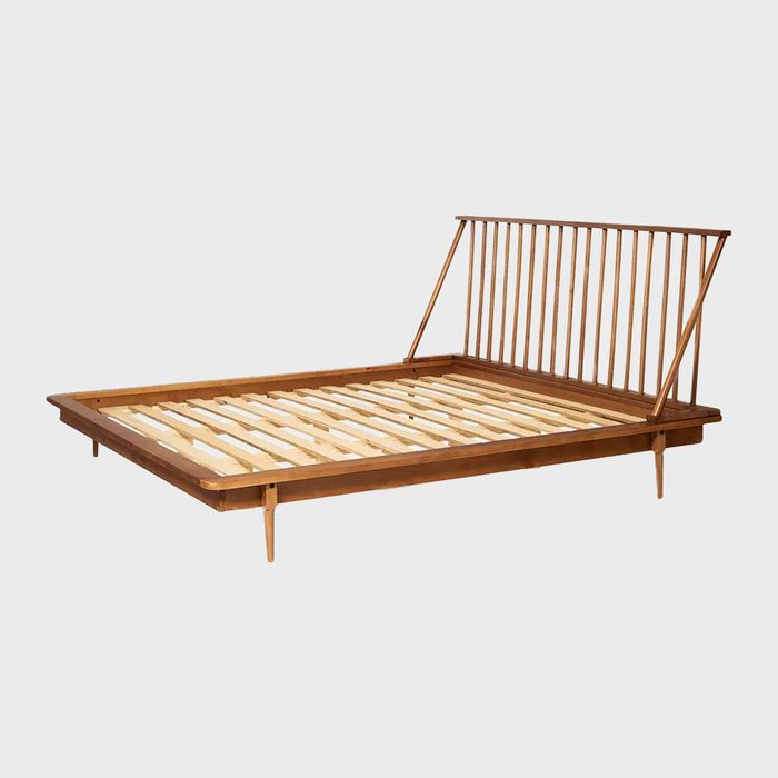 A wooden bed with a slatted base and an angled, spindle-backed headboard stands prominently against a light gray background.