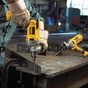 A worker uses a yellow DEWALT drill to bore into a metal surface on a workshop table, surrounded by tools in a well-lit industrial environment.