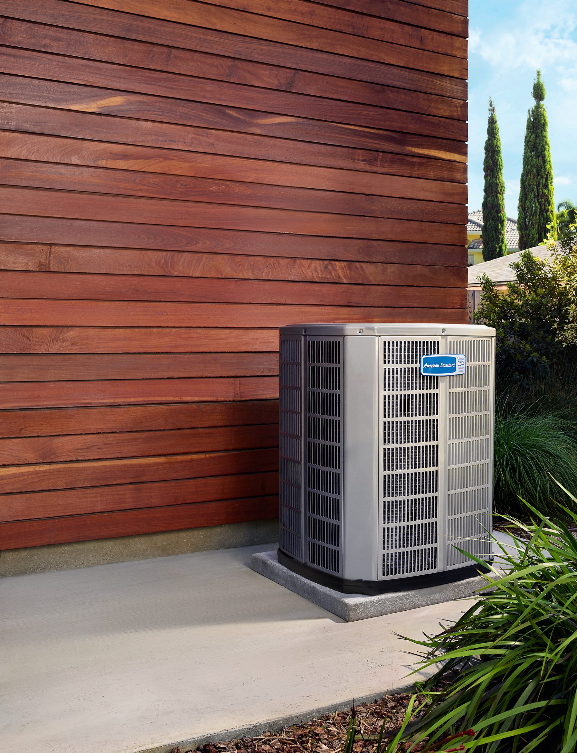 An air conditioning unit stands on a concrete pad beside a wooden wall, surrounded by plants and trees in a residential outdoor setting.