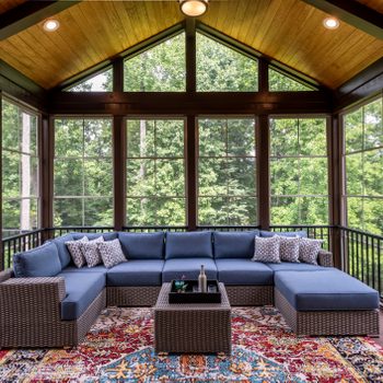 A spacious seating area features a sectional sofa and decorative pillows on a colorful rug, surrounded by large windows showcasing a lush green landscape.