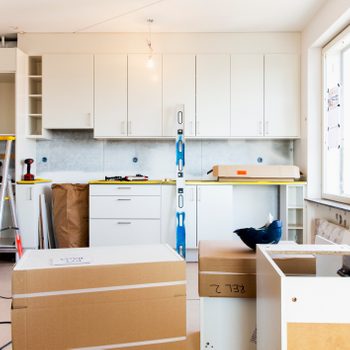 A ladder stands beside white cabinets being installed in a bright kitchen, with cardboard boxes and tools scattered across the unfinished floor.