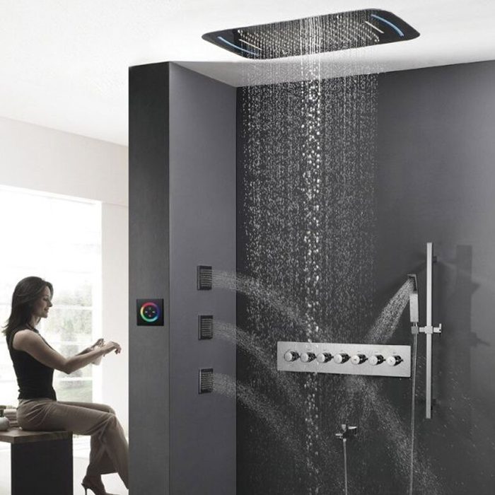 A modern shower system releases multiple streams of water, while a woman sits calmly, adjusting a digital control panel in a sleek bathroom environment.