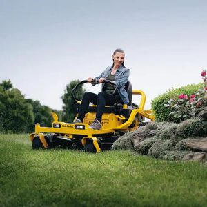 A woman operates a yellow riding lawnmower, cutting grass near a landscaped area with flowers and rocks, under a clear sky in a garden setting.