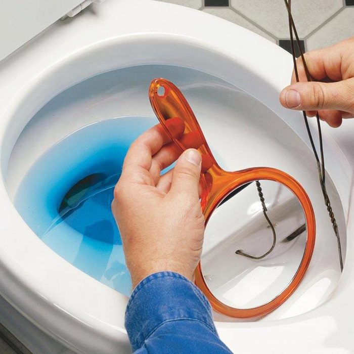 A hand holds an orange toilet auger above a blue liquid-filled toilet bowl, preparing to remove an obstruction in the plumbing.