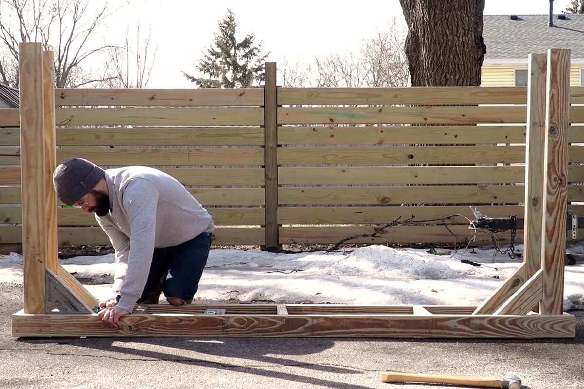 A man kneels to assemble a wooden frame in a snowy outdoor area, surrounded by a wooden fence and trees under a clear sky.