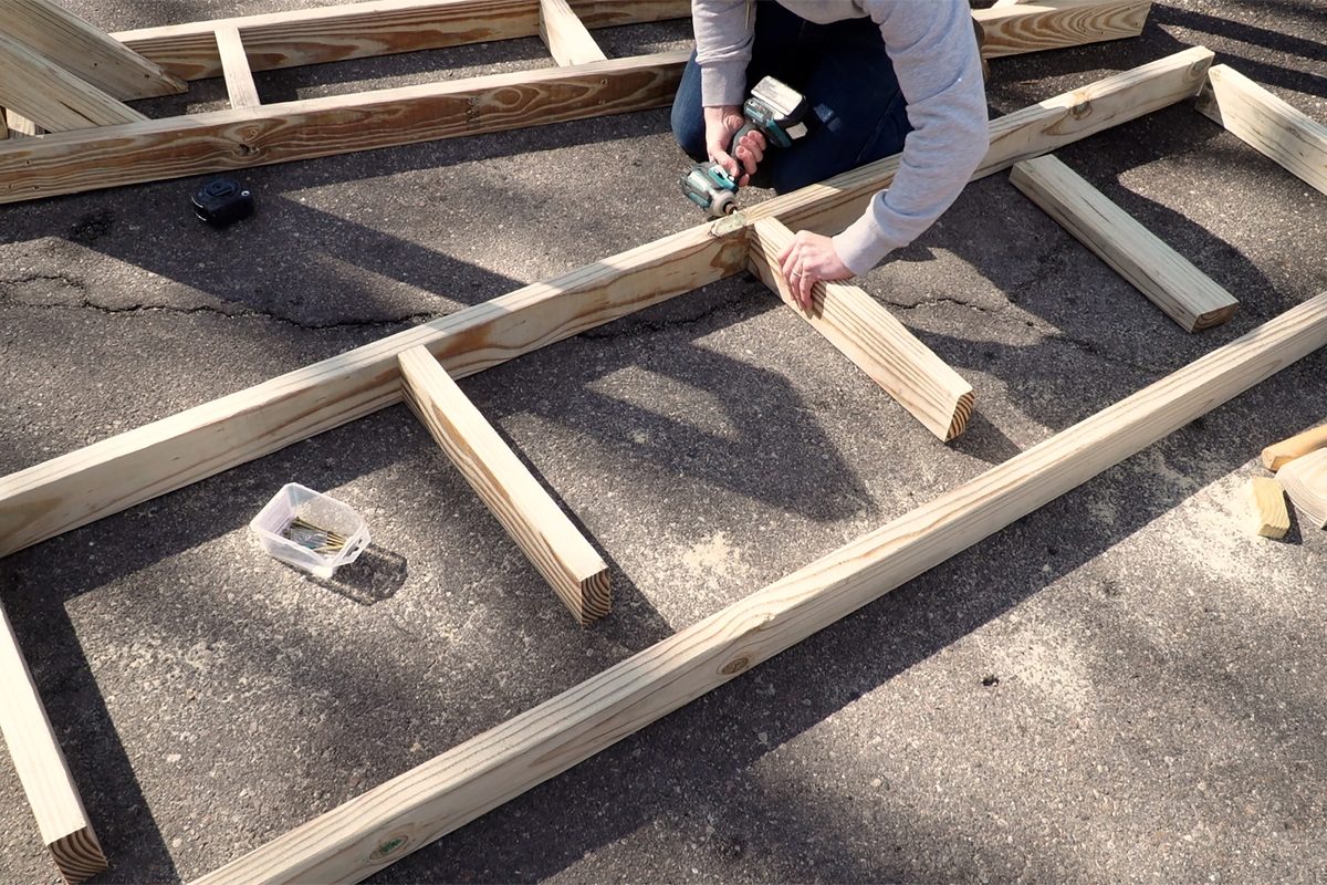 A person is screwing wooden planks together on a pavement, using a drill, with a small container of screws nearby.