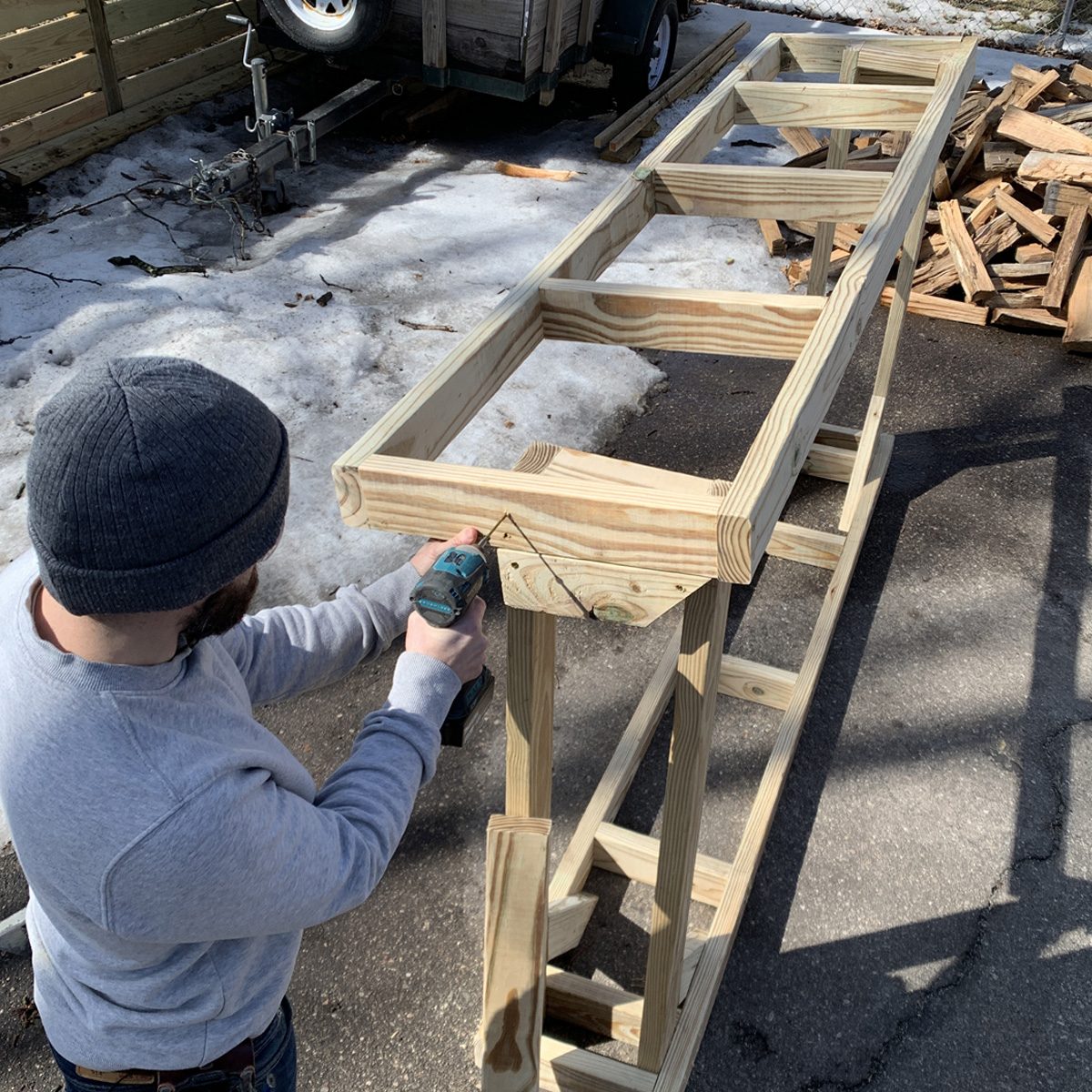 A person is using a power drill to work on a wooden frame outside, with snowy ground and construction materials in the background.