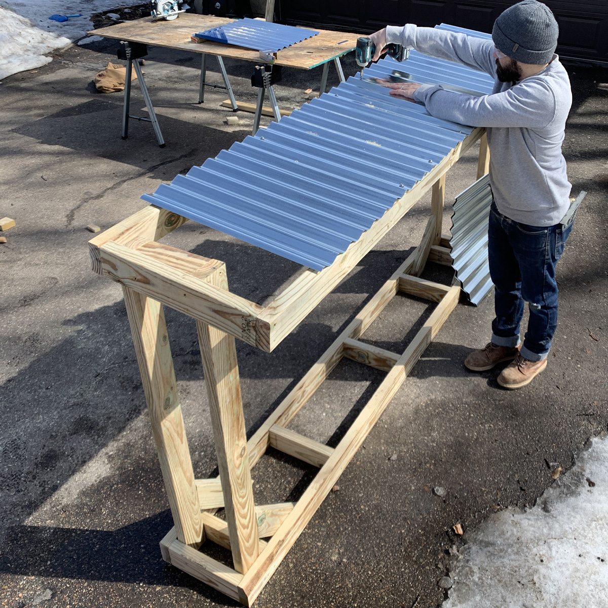 A person secures metal roofing sheets to a wooden frame using a power tool, on a sunlit driveway with construction materials nearby and snow in the background.
