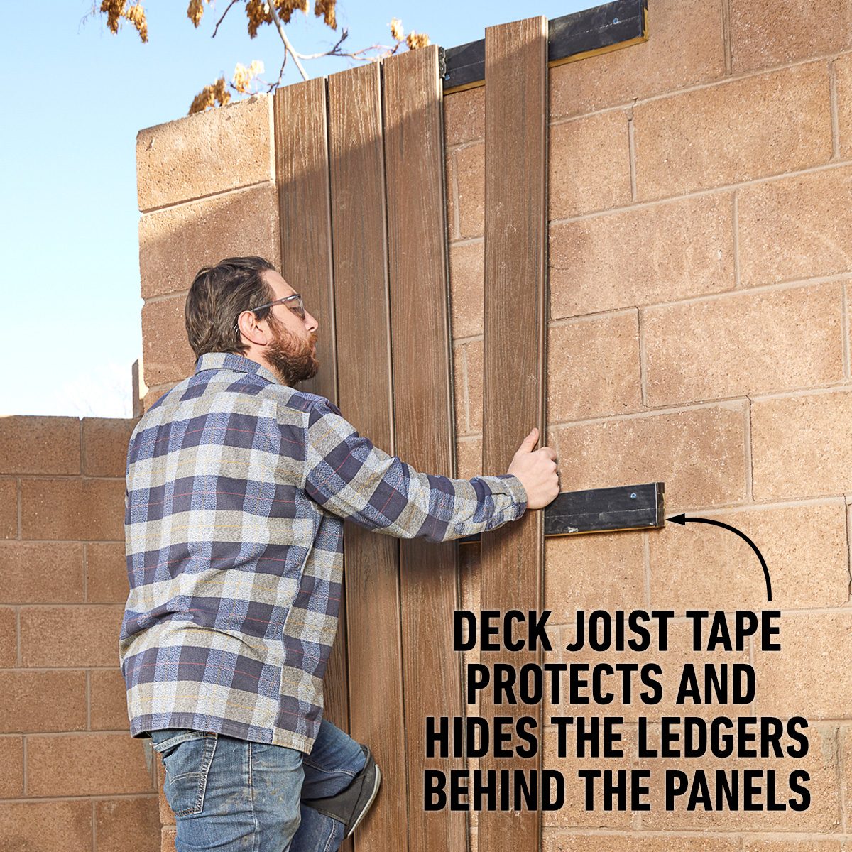 A man installs wooden panels onto a wall, securing them with tape, in a sunny outdoor environment featuring a tan brick wall.