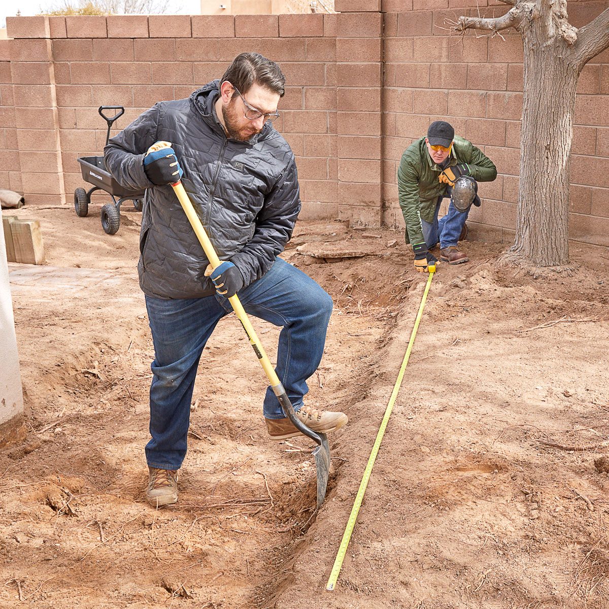 Two men are working in a dry, sandy yard; one digs with a shovel while the other measures a straight line with a tape measure.