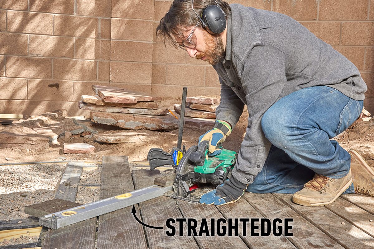 A person uses a power saw on wooden boards positioned over gravel, ensuring precise cuts with a straightedge tool in a construction environment.