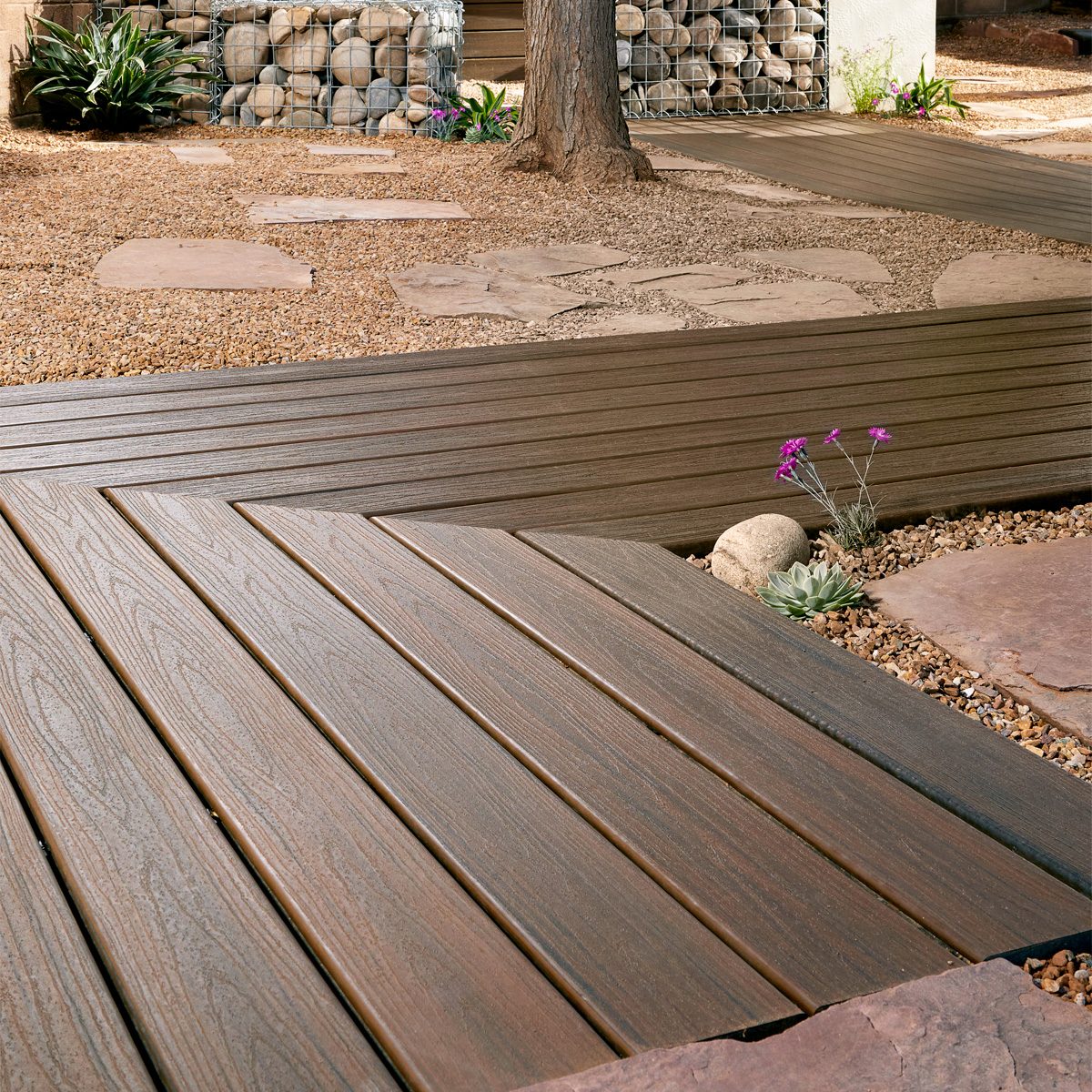 Wooden decking boards create a pathway, intersecting in a zigzag pattern across a gravel landscape, surrounded by stone accents and small plants.