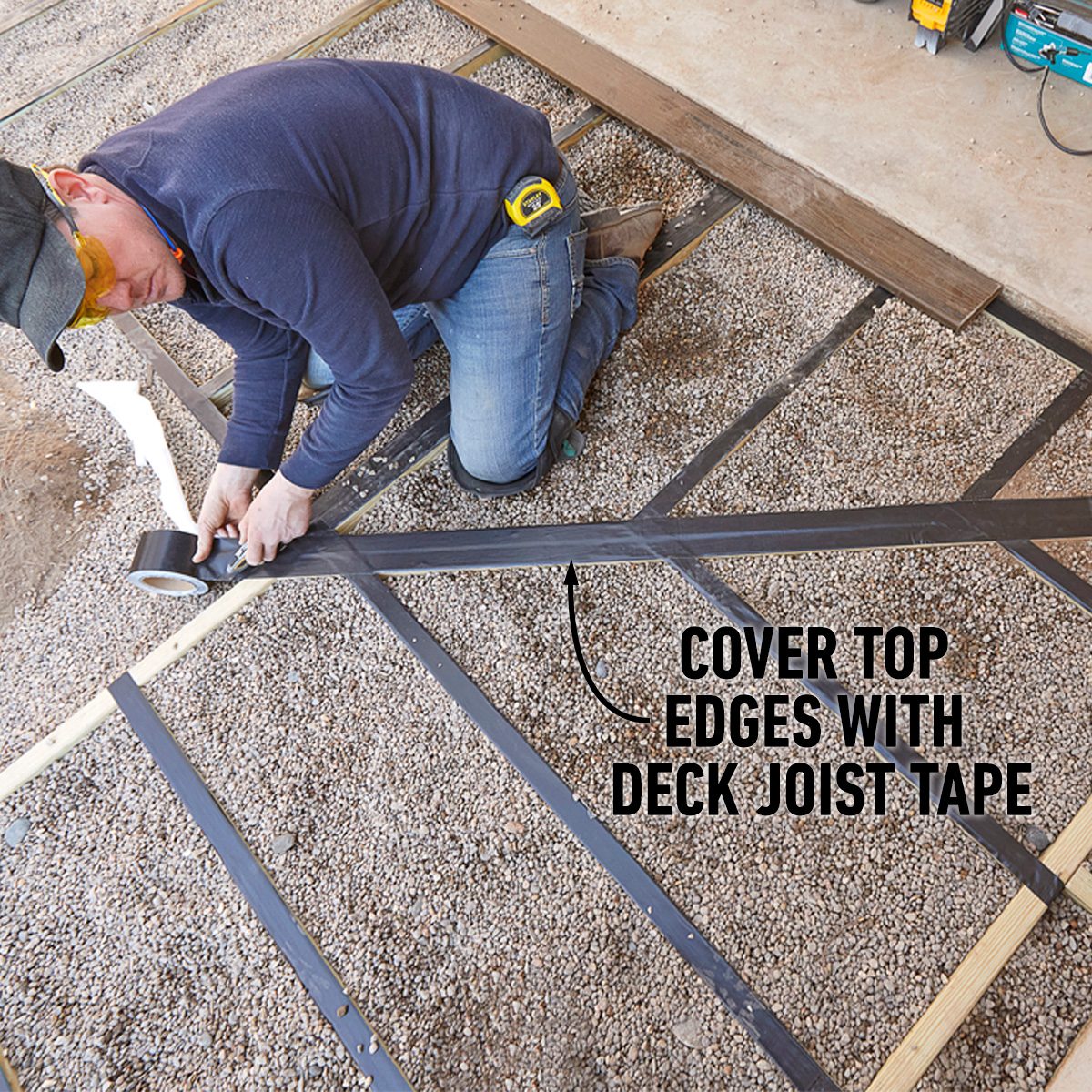 A person kneels on gravel, applying deck joist tape to wooden beams, while using a tape measure. Sunlight illuminates their work area.