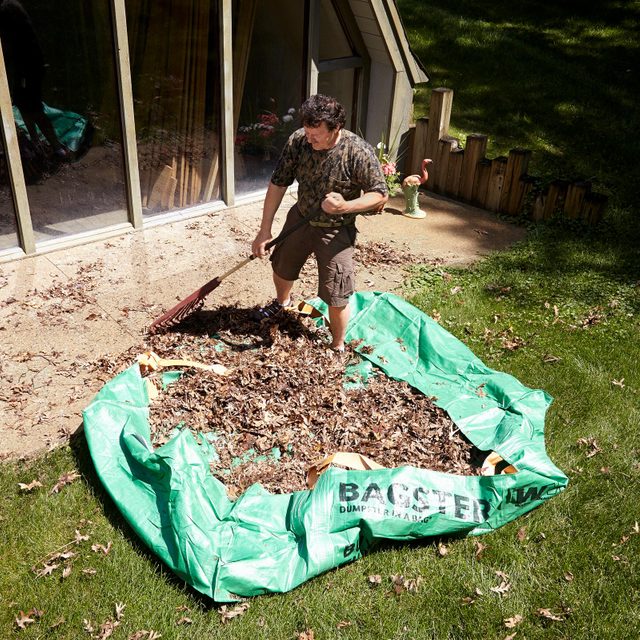 A man rakes leaves into a large green bag in a sunny backyard, near a house with large windows and decorative items.