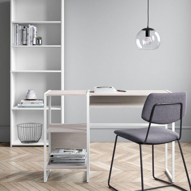 A modern desk with a gray chair sits on a herringbone floor, illuminated by a pendant light, next to a white bookshelf filled with books.
