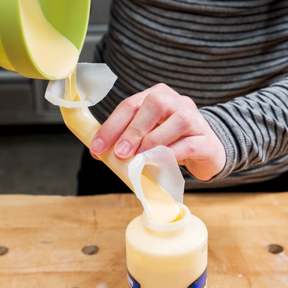 A hand pours a creamy substance from a green container, directing it into a bottle while holding a paper towel in place on the bottle's opening.