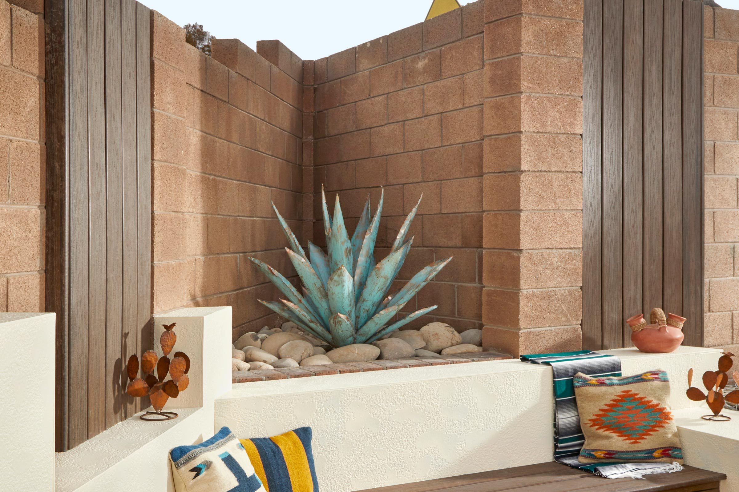 A large blue agave plant sits among smooth stones in a cozy outdoor nook, surrounded by tan brick walls and adorned with decor and textiles.