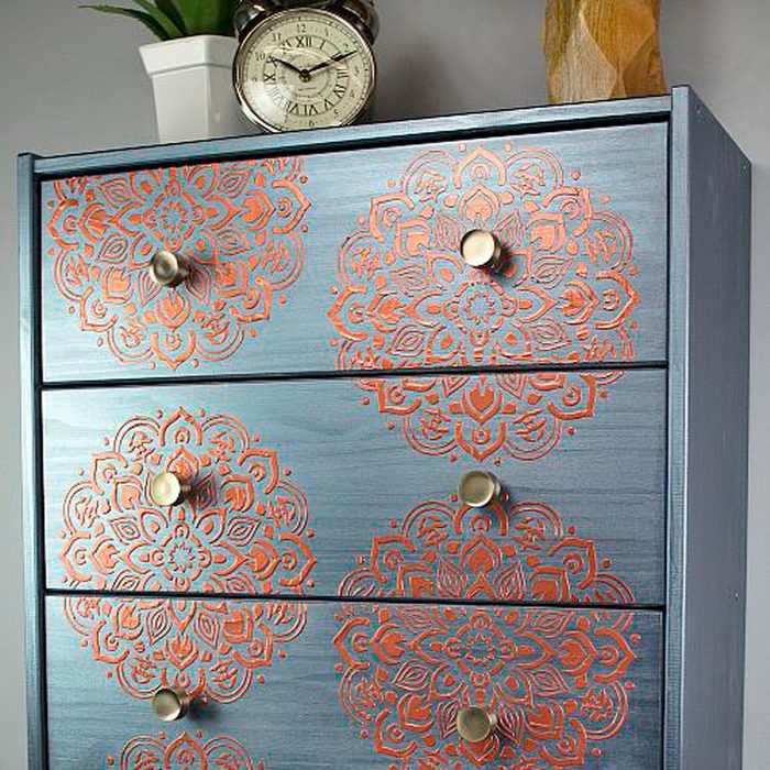 A blue dresser features intricate orange mandala designs on its front. It's accented by round gold knobs and topped with a clock and potted plant.