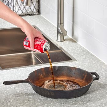 A hand pours soda from a can into a cast iron skillet on a speckled countertop near a sink.