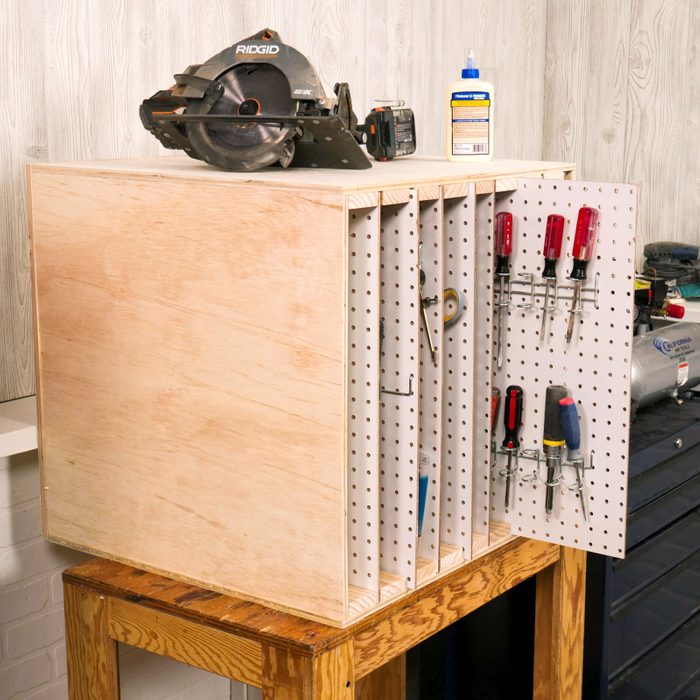 A wooden tool storage unit displays various tools on pegboard, with a circular saw, battery, and glue bottle placed on top against a light-colored wall.