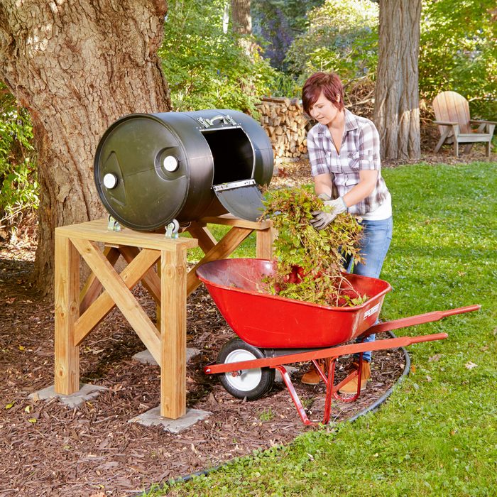 A person discards plant waste into a black compost barrel supported by wooden legs, while a red wheelbarrow sits nearby on grass.