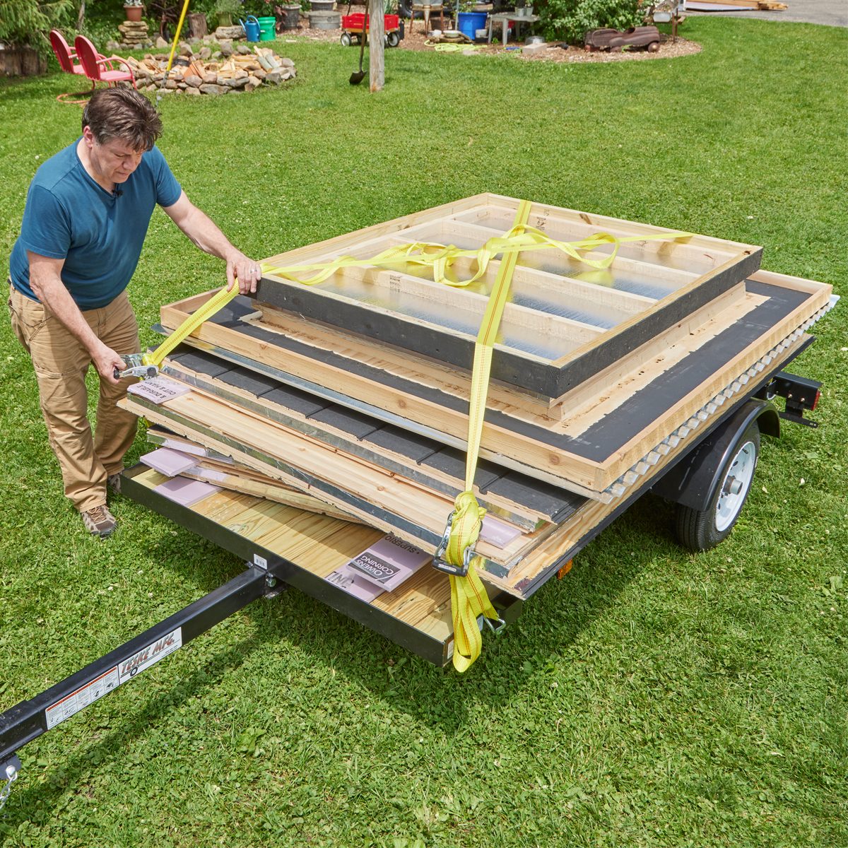 A man secures building materials on a trailer with straps, surrounded by green grass and various outdoor objects in the background.