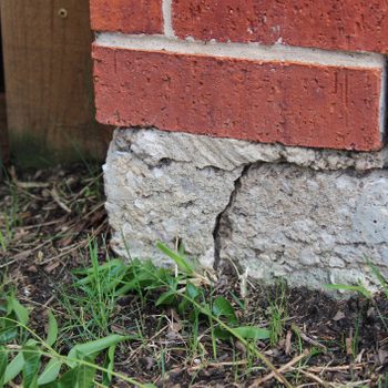 A brick wall stands above a cracked concrete base, surrounded by damp soil and patches of green grass, with a metal pole nearby.