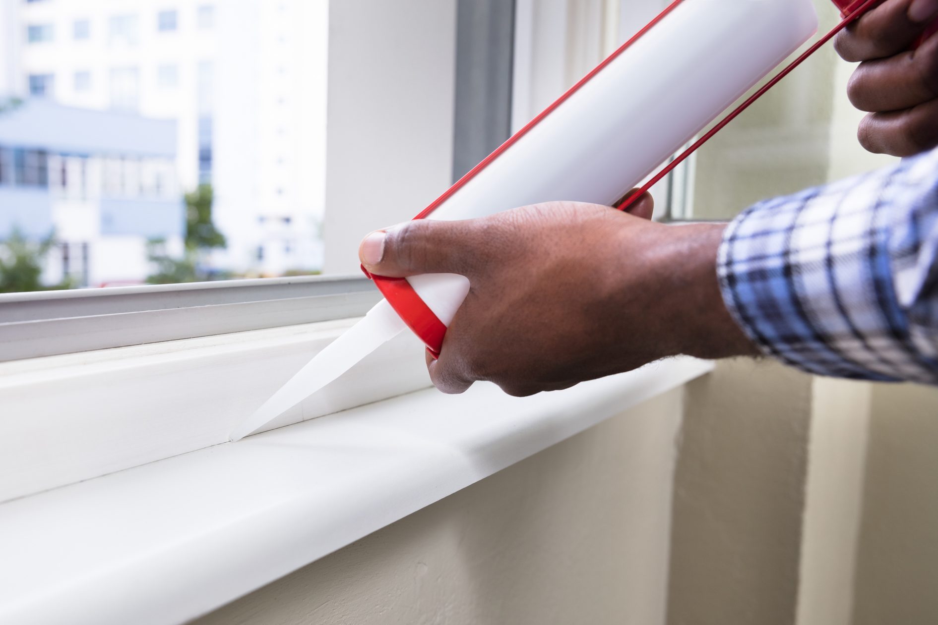 A person applies caulk from a cartridge along a window sill, with buildings and greenery visible through the window.