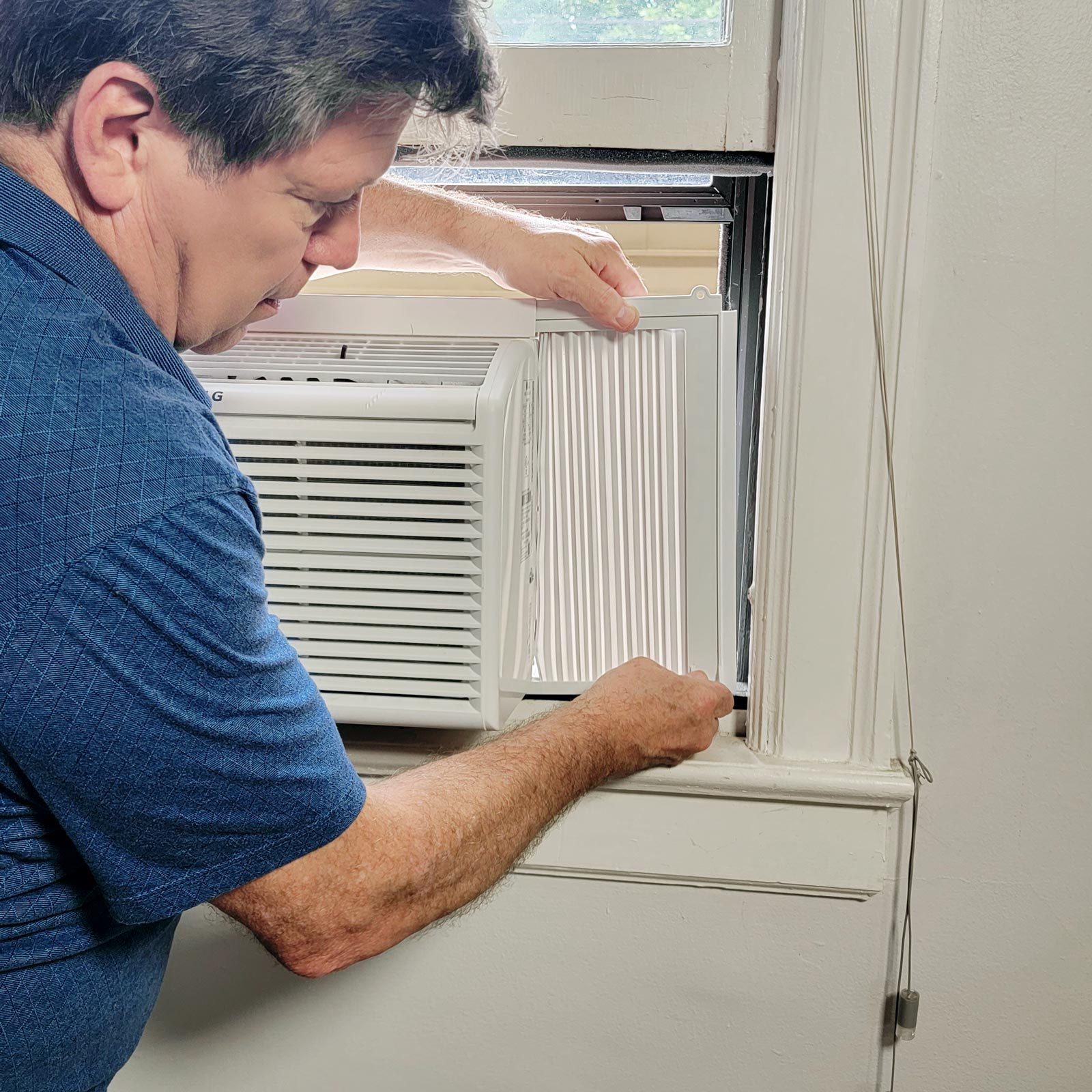 A man removes a filter from a window air conditioner, positioned in a white-painted window frame with natural light illuminating the scene.