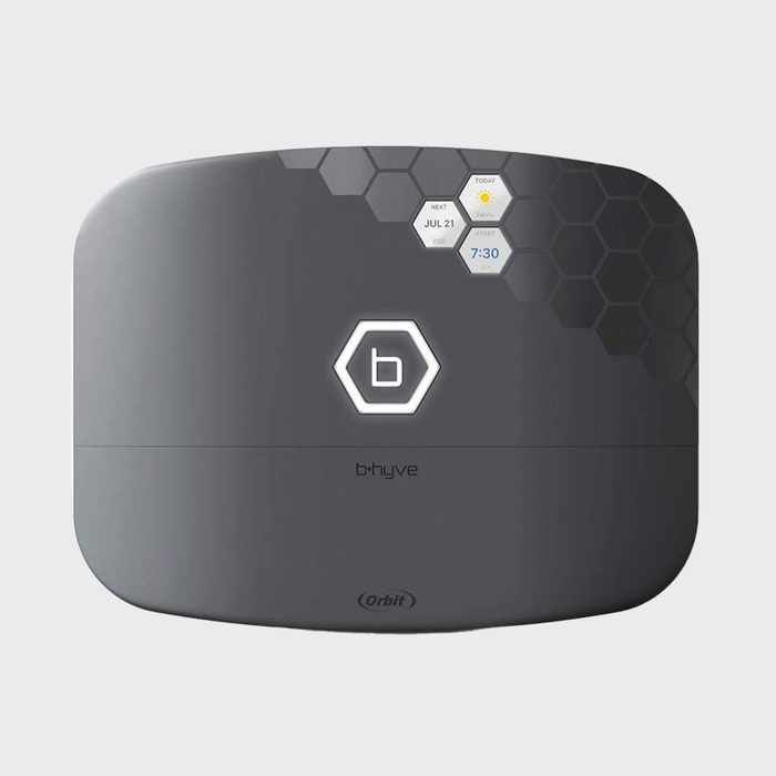 A gray smart irrigation controller displays a digital interface showing date, weather, and schedule settings against a neutral background.