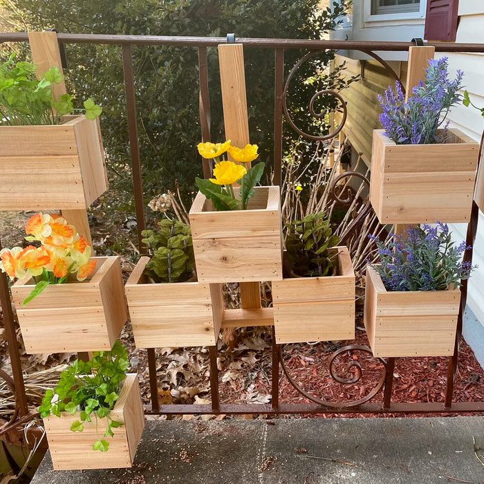 Wooden planter boxes display colorful flowers and plants, hanging on a railing against a backdrop of greenery and mulch on the ground.