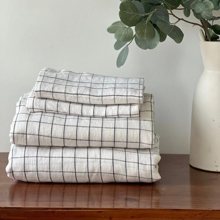 A stack of folded, grid-patterned linens rests on a wooden surface, accompanied by a vase with green leaves against a light wall.