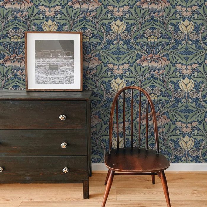A wooden chair sits beside a dark dresser, adorned with a framed photo, against a floral-patterned wallpaper in a well-lit room.