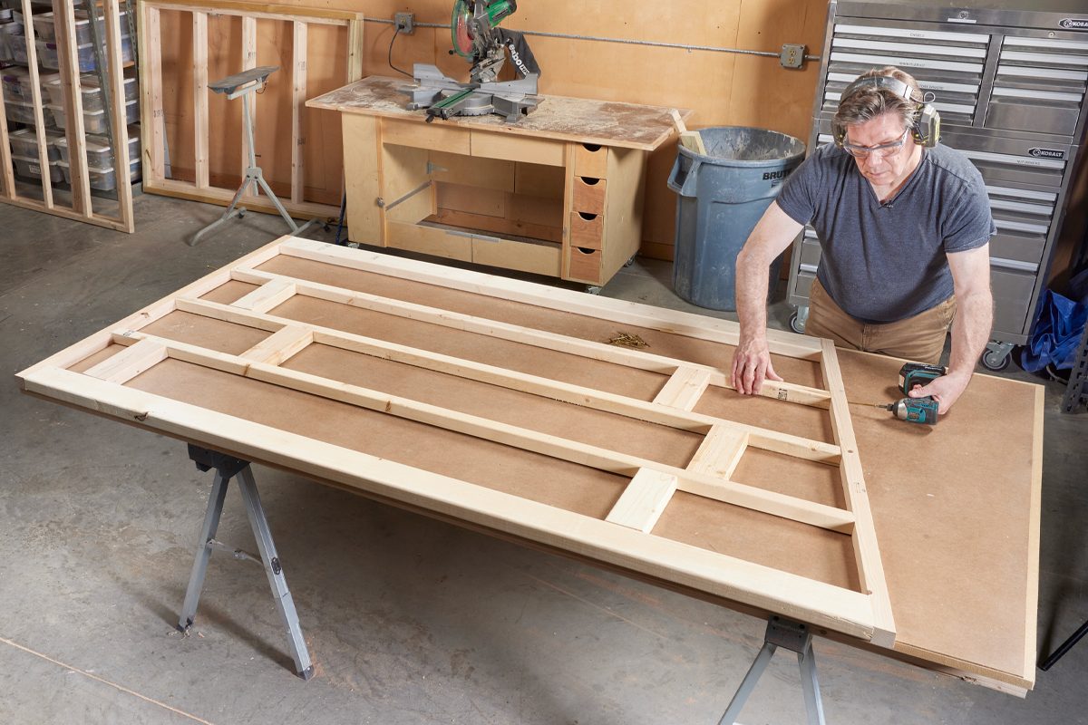 A person uses a drill to assemble a wooden frame on a worktable in a workshop, surrounded by tools and partially completed projects.