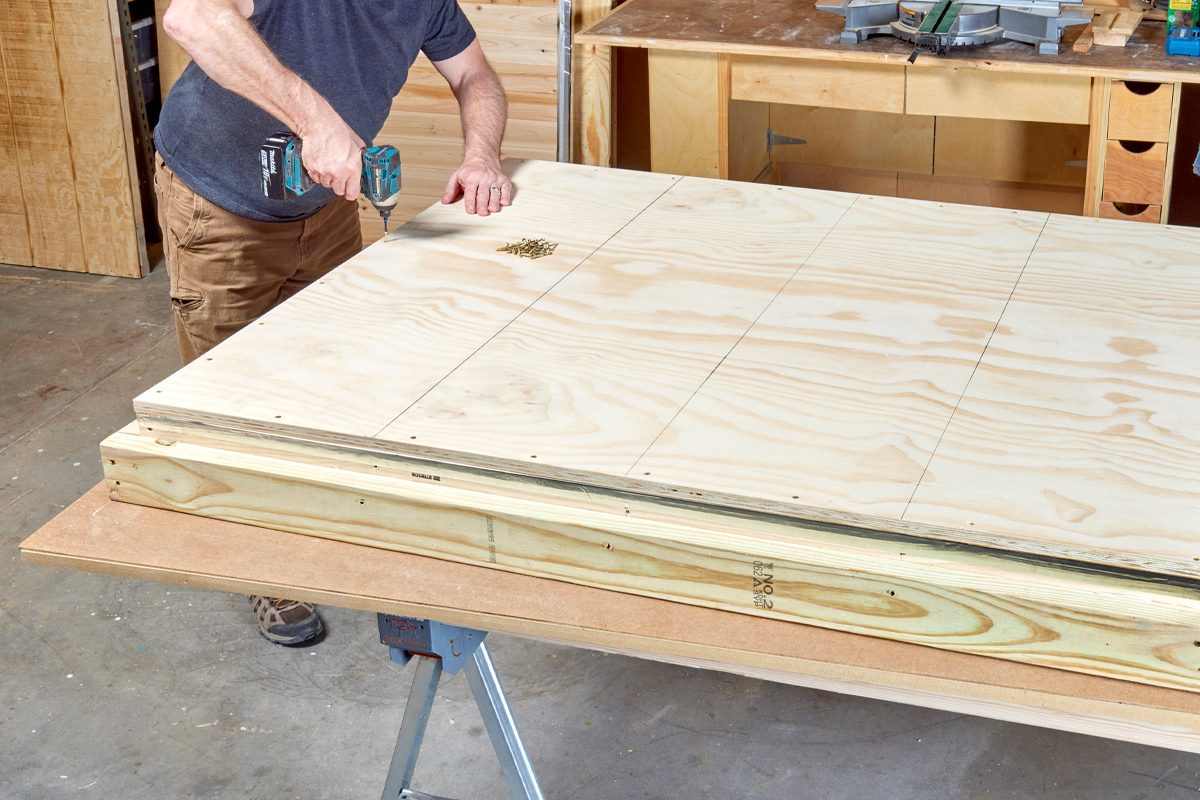 A person drills into a large wooden panel on a workbench, surrounded by tools and materials in a workshop.