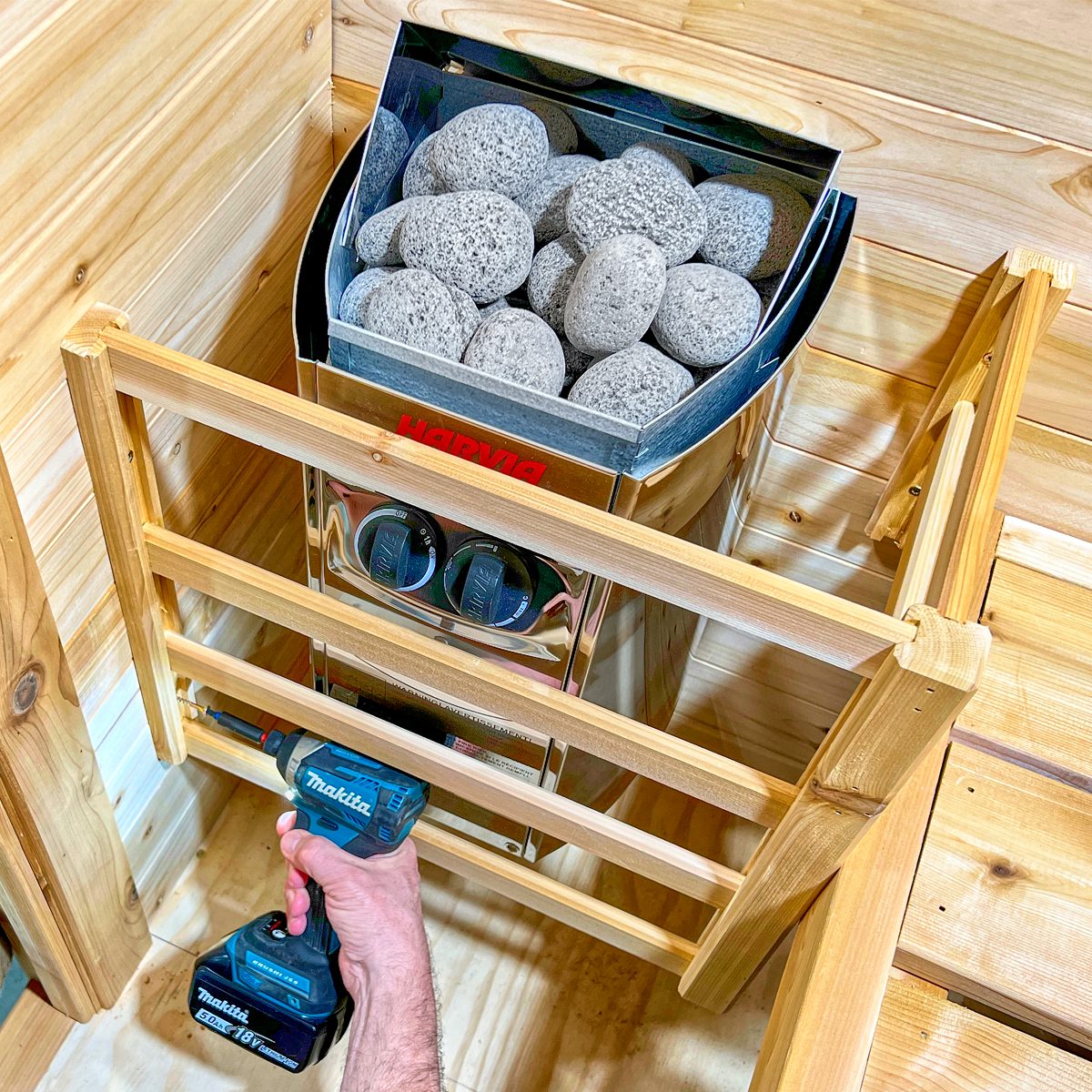 A hand operates a drill, assembling wooden slats around a heater filled with gray stones, situated in a wooden interior space.
