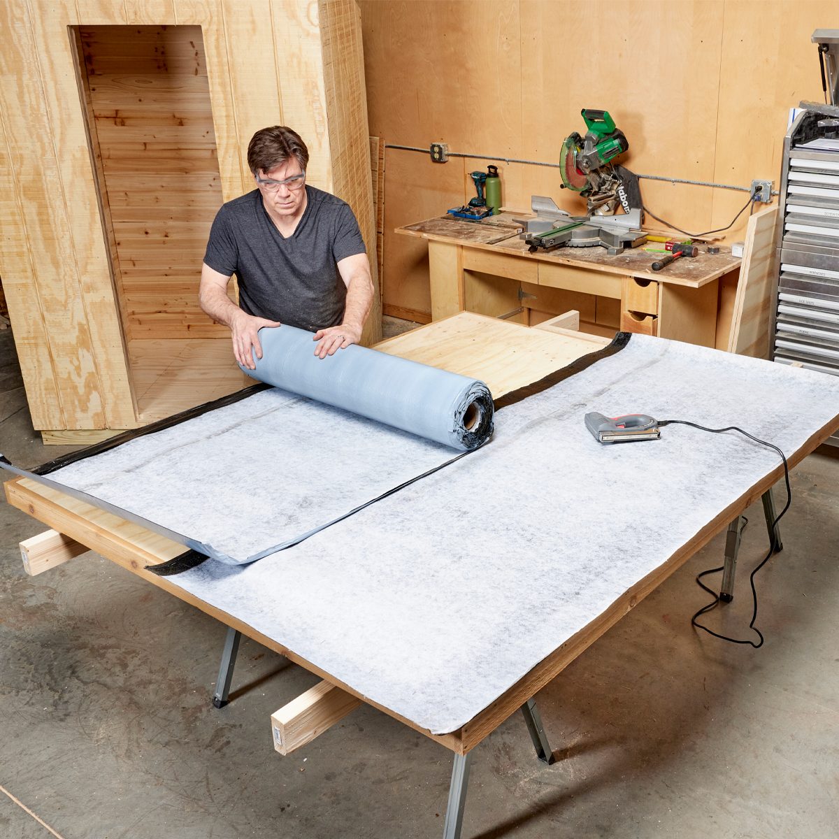 A man unrolls a blue fabric roll onto a table in a workshop, surrounded by wooden structures and tools, preparing the material for a project.