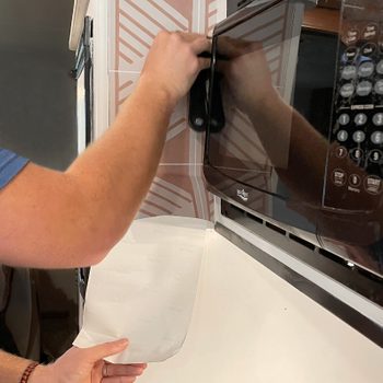 A person holds a piece of paper while adjusting a microwave's installation against a patterned wall in a kitchen setting.